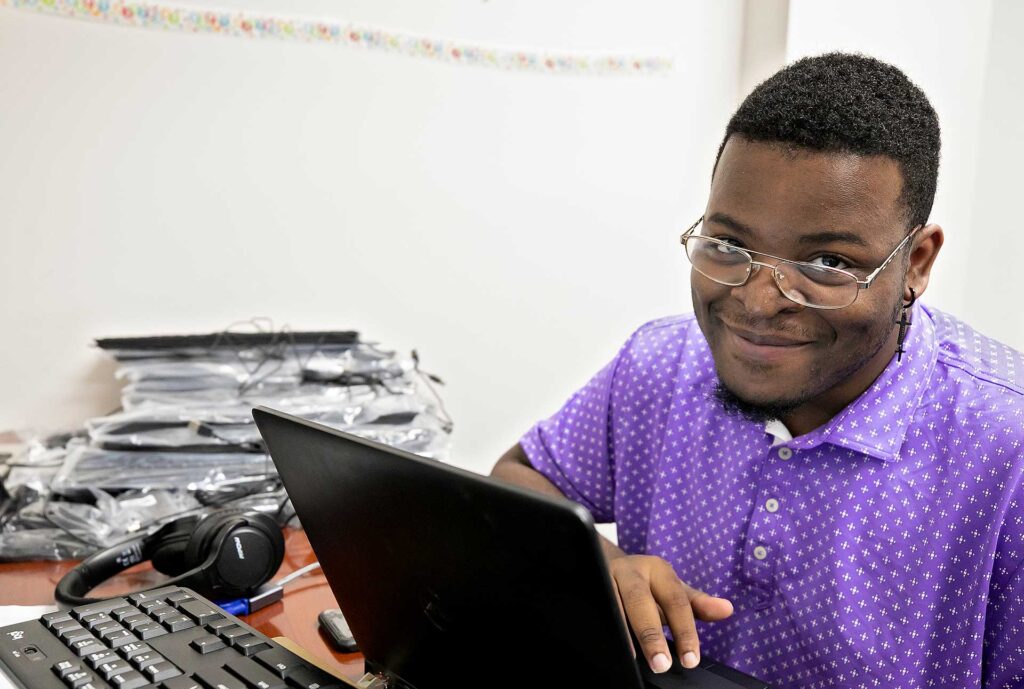 A young man smiles while working on a laptop
