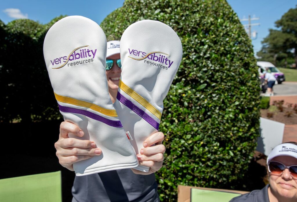 A person smiling while holding up golf club head covers with VersAbility logo on them