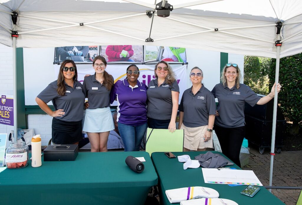 A group of women in golf attire smile and pose from behind a table under a tent