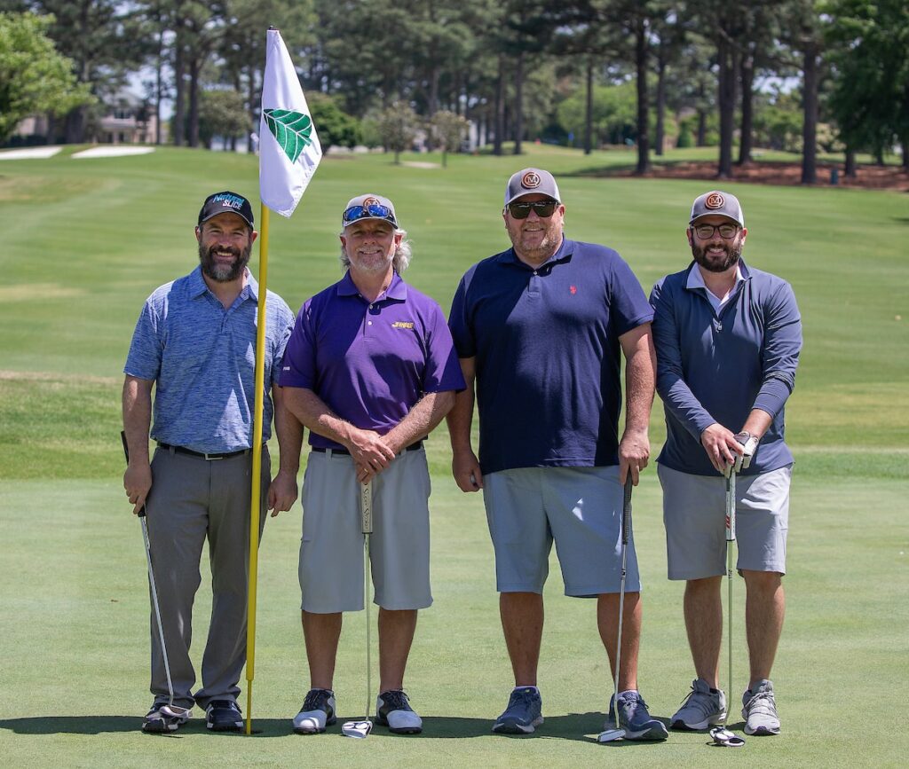 Four men in golf attire pose with their golf clubs near a flagstick on a golf course