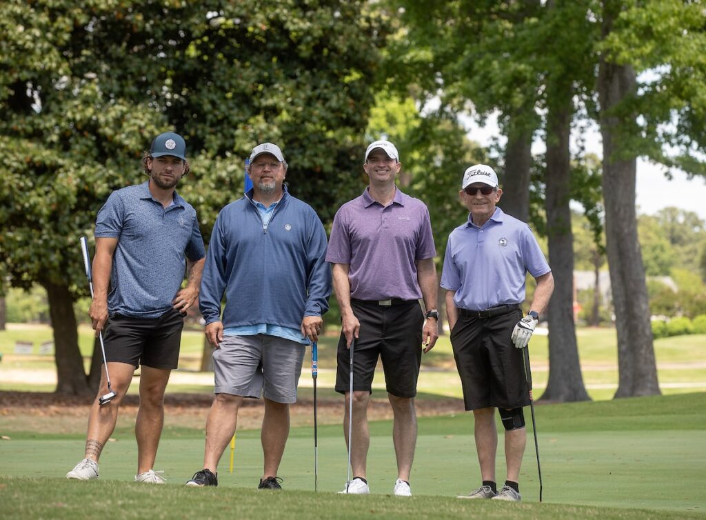 A group of four men in golf attire pose with their golf clubs on a golf course