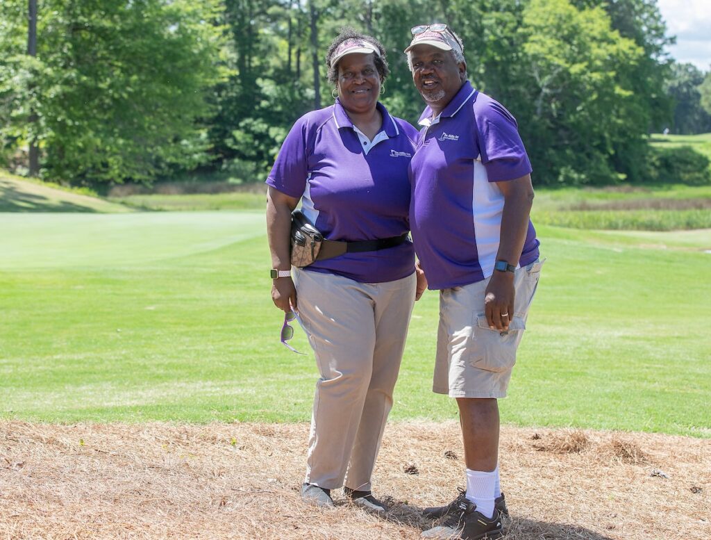 Two people in purple golf shirts stand next to each other and smile on a golf course