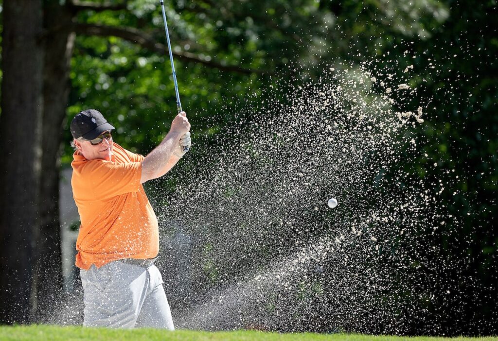 A man in an orange golf shirt hits a golf ball with his club on a golf course