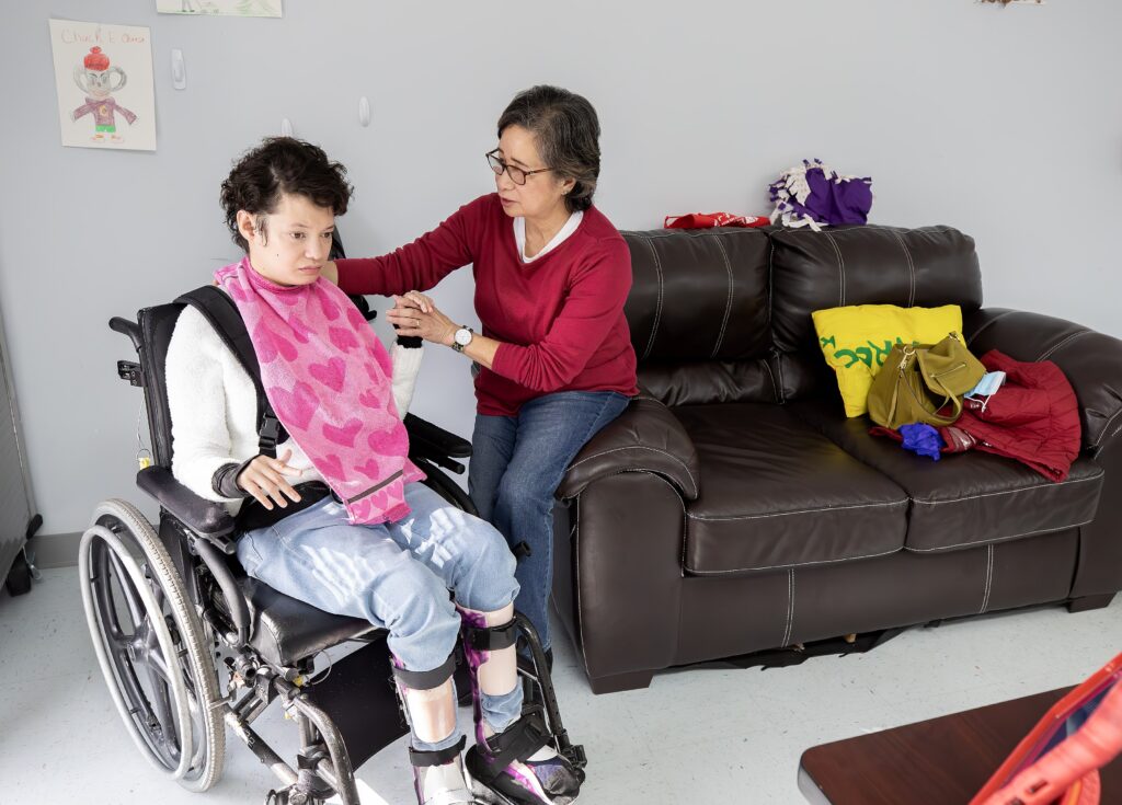 Mary Jane Teall sits on the arm of a couch while holding the hand of her daughter, Lauren, who is seated next to her in a wheelchair