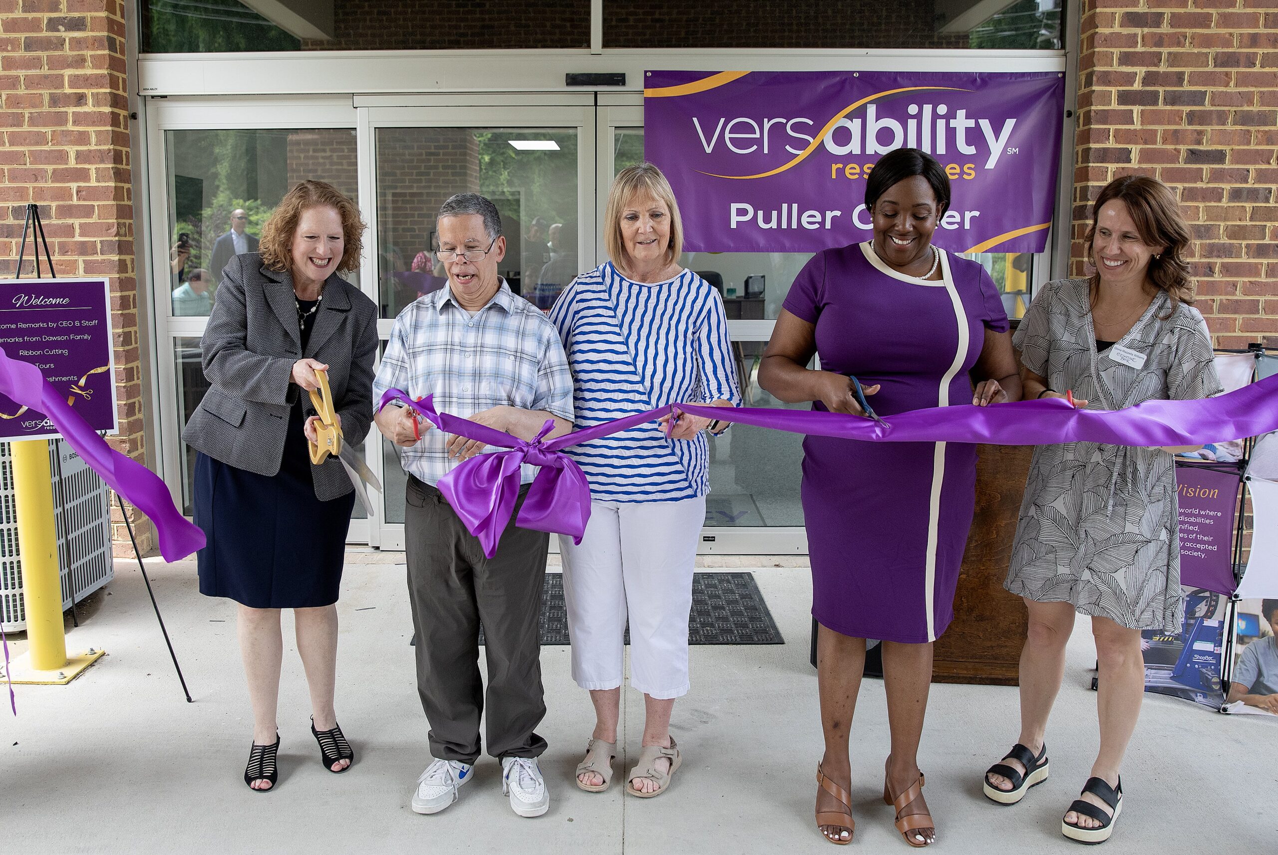 Four women and one man stand in front of the Puller Center while one of the woman cuts a large purple ribbon in front of the doors