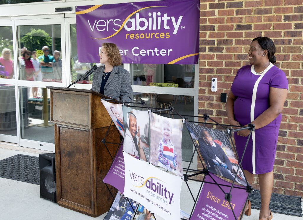 A woman speaks at a podium in front of the Puller Center while another woman in a purple dress stands and listens nearby
