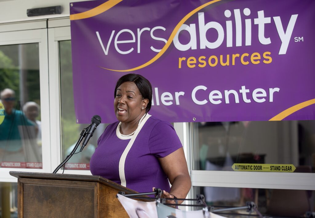 A woman speaks into a microphone at a podium in front of a purple VersAbility banner