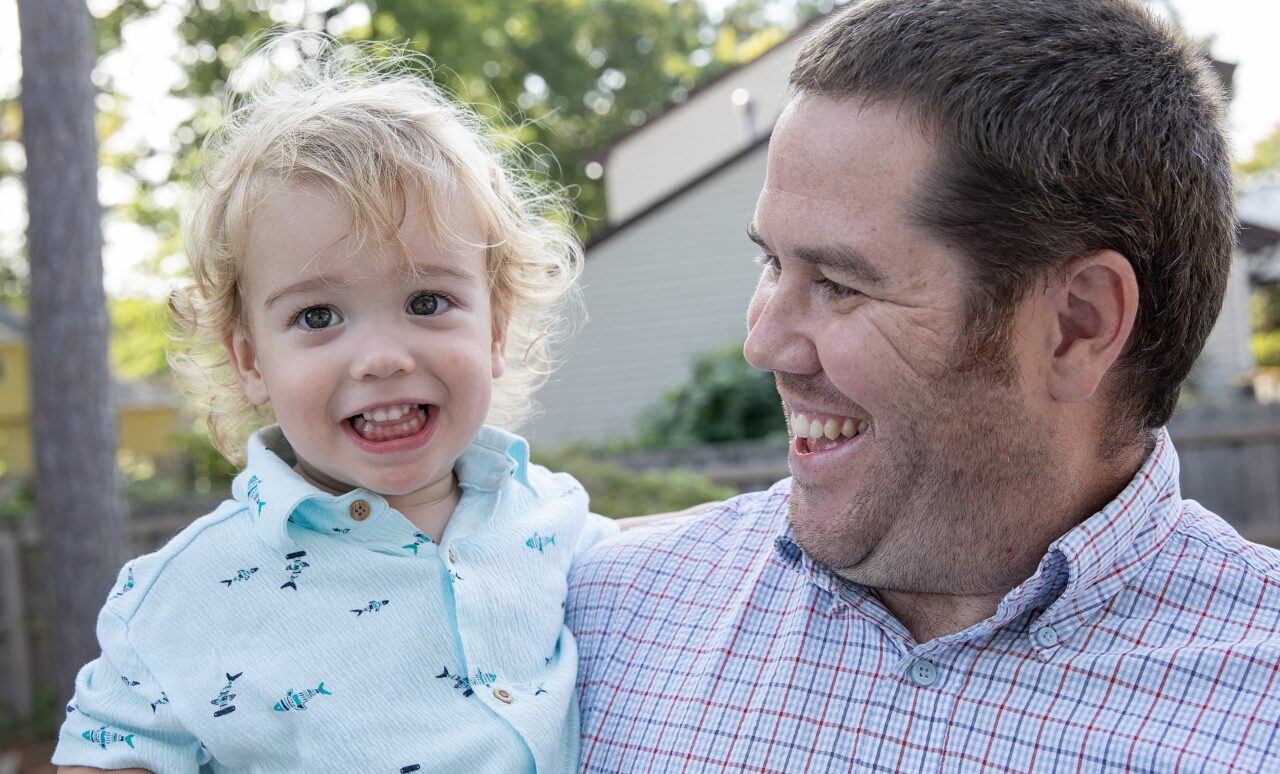 A smiling man holds a cheerful, curly-haired toddler outdoors. Both are looking at each other and smiling, radiating the joy and connection often fostered through Early Childhood Intervention, with trees and buildings blurred in the background.