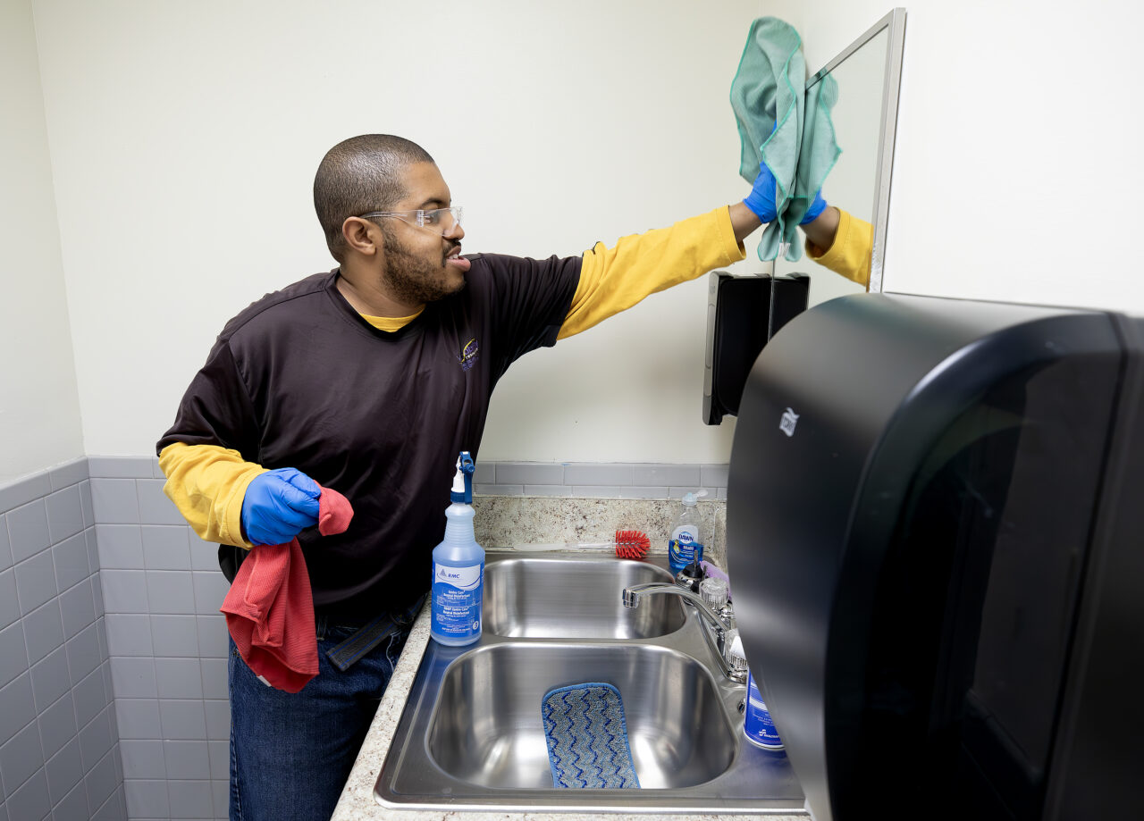 A VersAbility worker wearing gloves cleans a bathroom mirror near a sink, holding a cloth in one hand and a spray bottle on the counter. Paper towels and soap are visible nearby.
