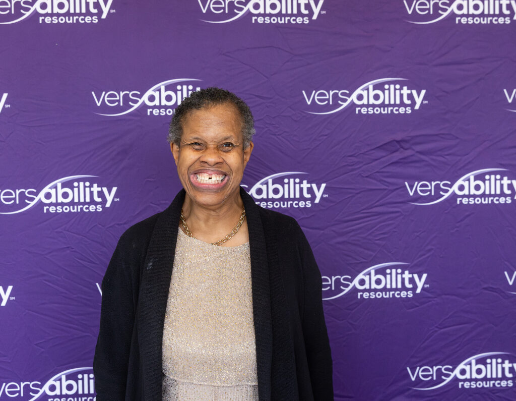 Woman in a gold top and black cardigan smiling for the camera in front of VersAbility Resources background.