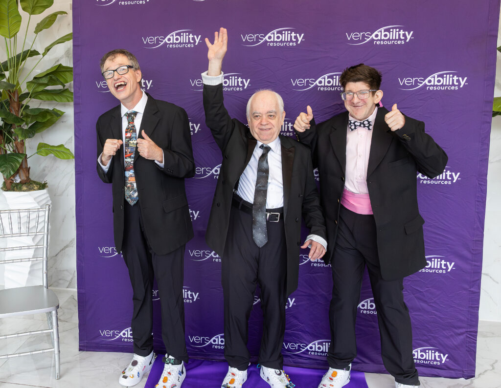 Three men in suits smile and pose in front of purple VersAbility Resources background
