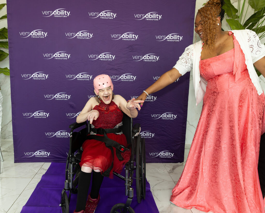 A woman in a red dress sits in a wheelchair, smiling and holding hands with a woman in a peach dress