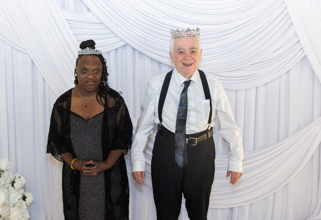 A woman in a dress wearing a tiara and man in a suit wearing a crown, standing next to each other, both smiling.