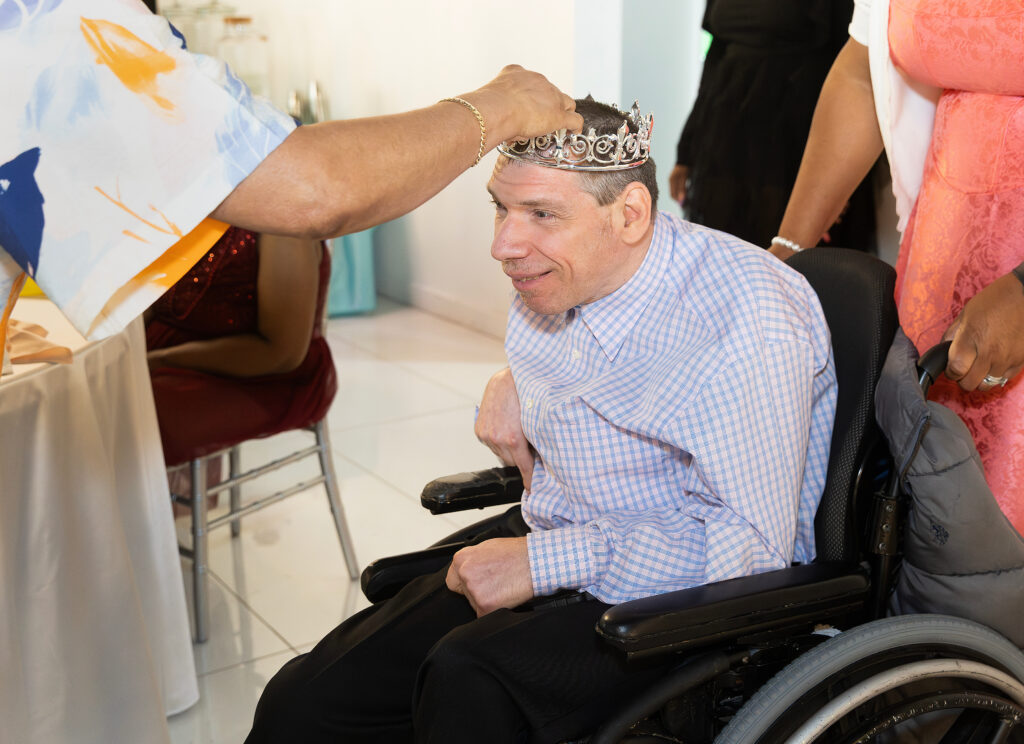 A man in a blue checkered shirt, sitting in a wheel chair smiles as he receives a crown 