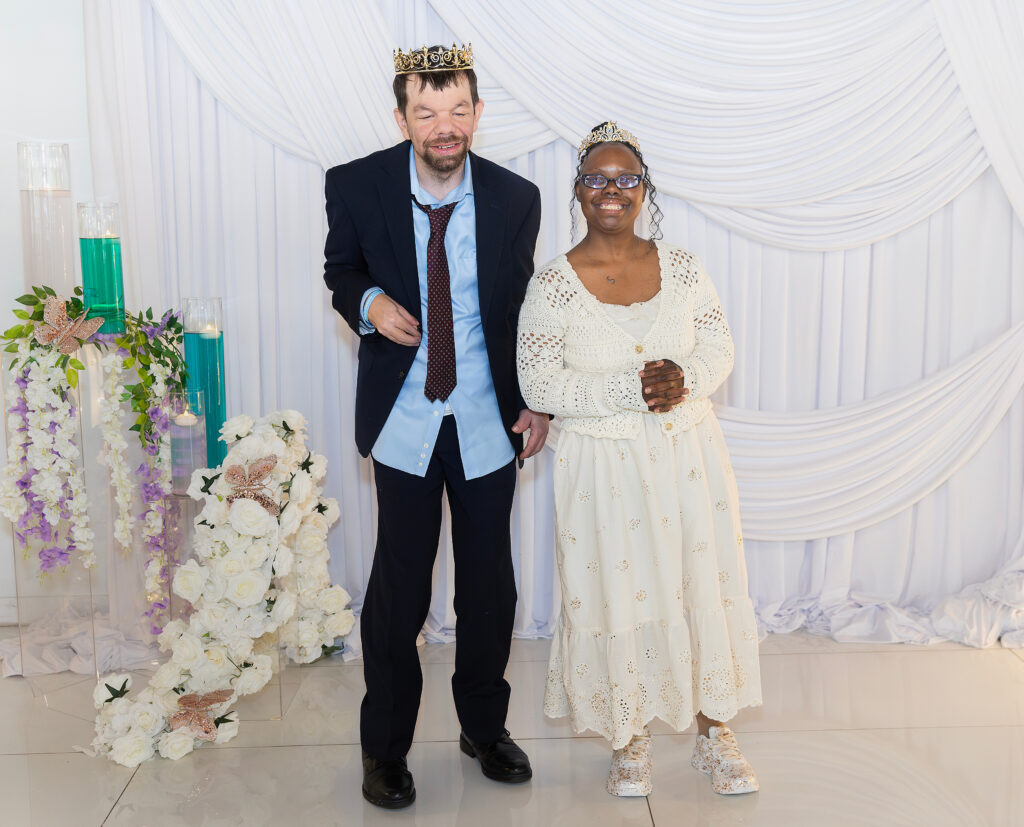 A woman in a dress wearing a tiara and man in a suit wearing a crown, standing next to each other, both smiling.