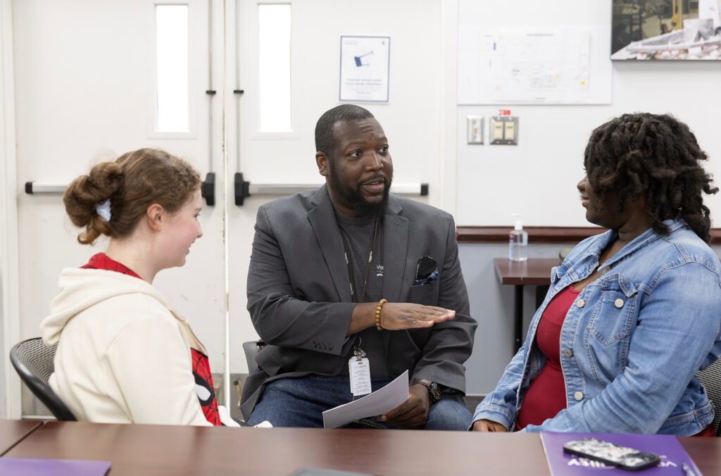 A man sits between two women at a table, engaging in conversation. He gestures with one hand while holding papers in the other.