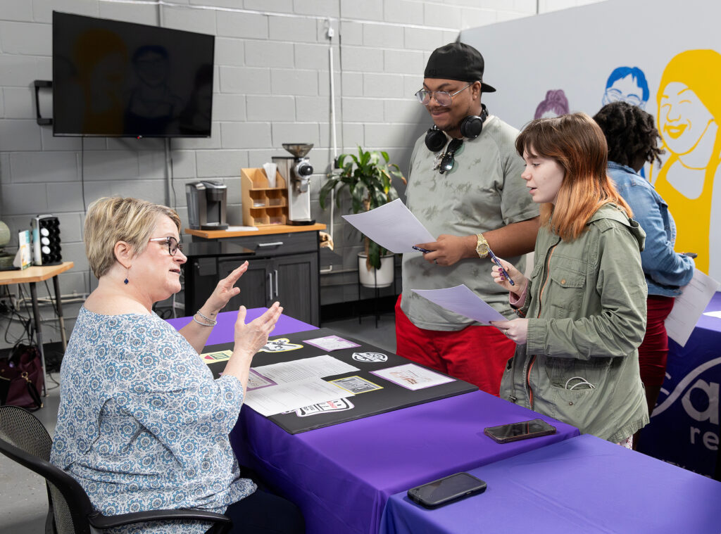 A woman at a table with informational brochures discusses with two young people holding papers.