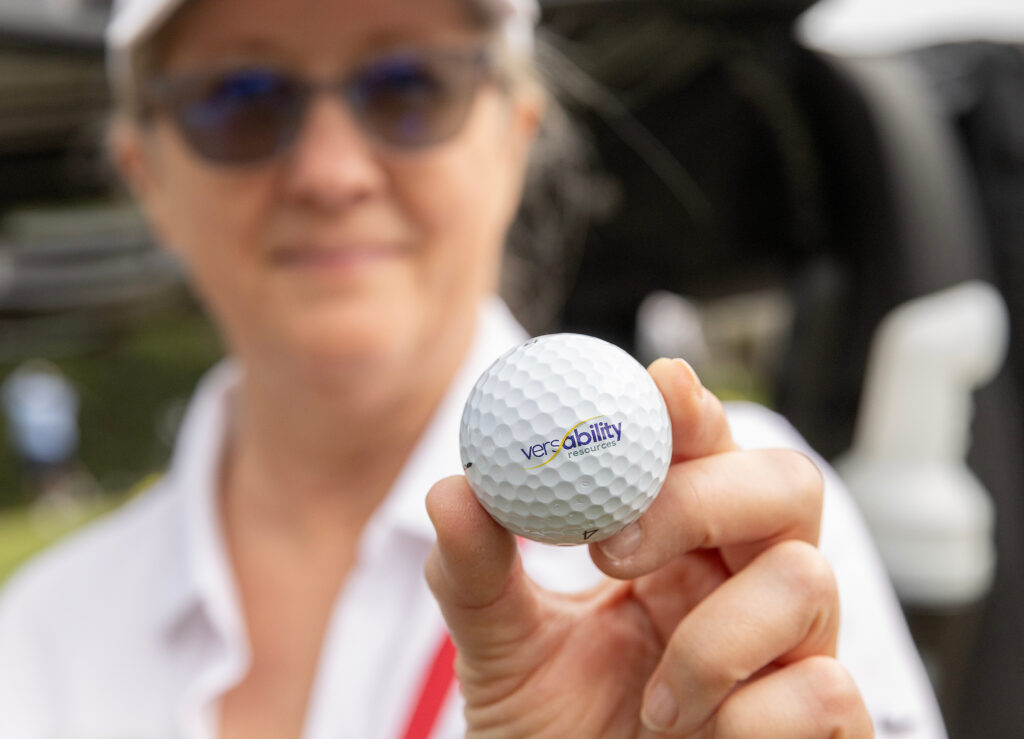A woman holds a golf ball with the VersAbility logo, highlighting the VersAbility golf tournament event.