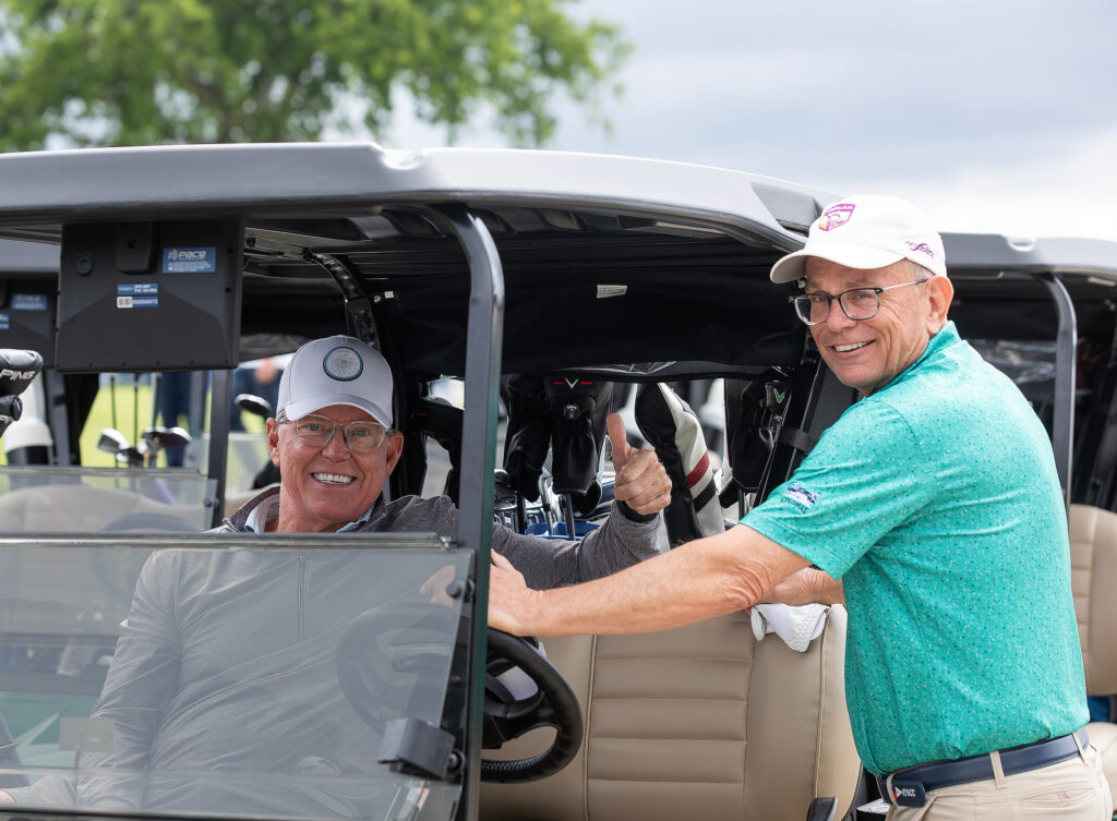 Two smiling men in a golf cart at the VersAbility golf tournament, one giving a thumbs-up, enjoying the event.