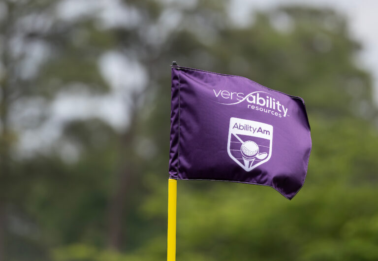 A purple flag with “VersAbility Resources” and “Ability Am” logos waves on a yellow pole, celebrating the VersAbility charity golf tournament, with blurred greenery in the background.
