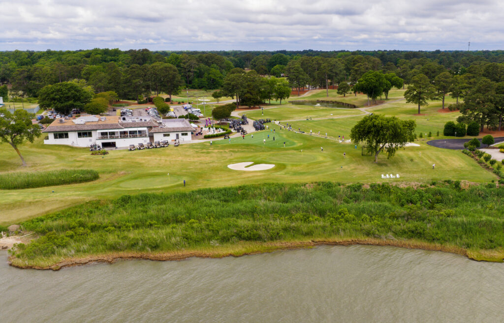 Wide show of a lush course by water during the VersAbility golf tournament, with a clubhouse and parked carts visible nearby.