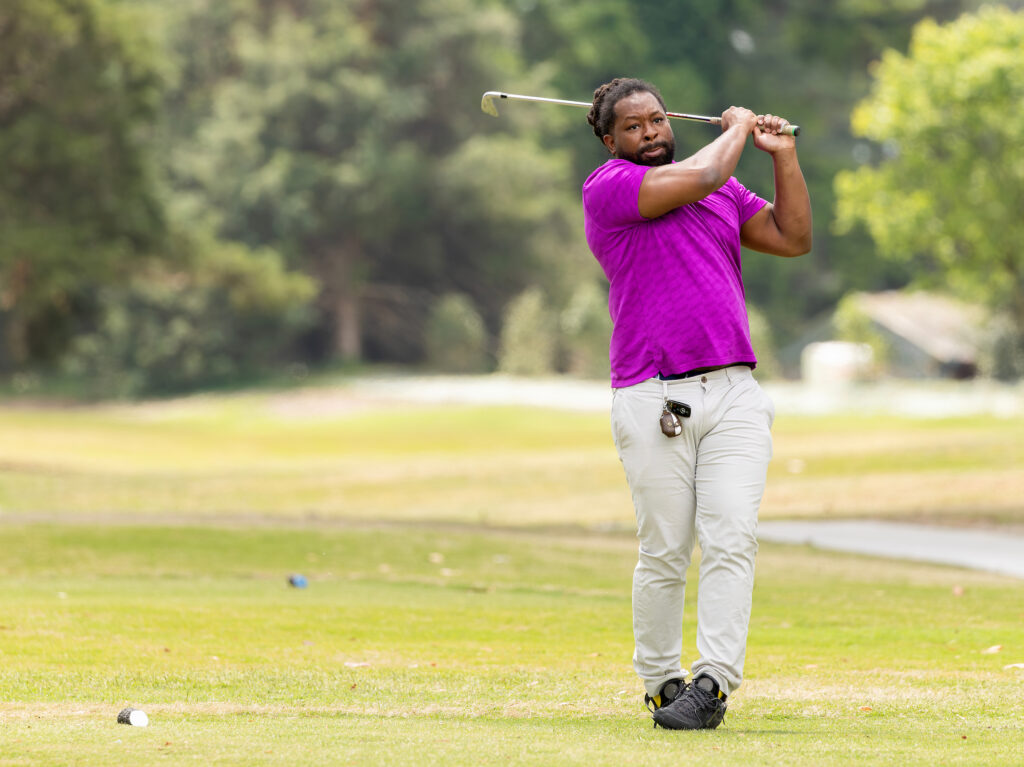 Golfer in purple shirt swings at VersAbility golf tournament on a sunny course, surrounded by lush green trees.