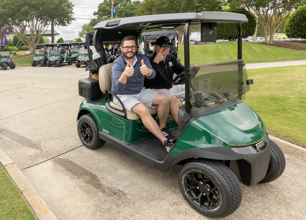 Two men in a golf cart give thumbs up at the VersAbility golf tournament, surrounded by lush greenery and other carts.