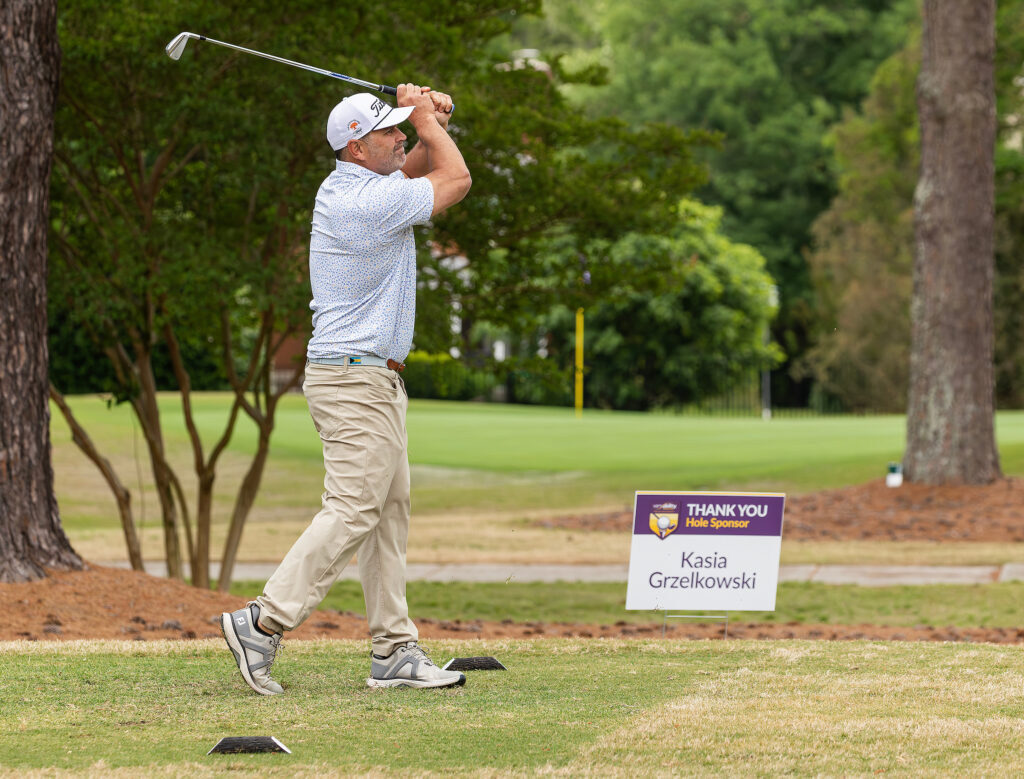 Golfer swings at the VersAbility golf tournament, surrounded by trees and a sponsor sign on the course.