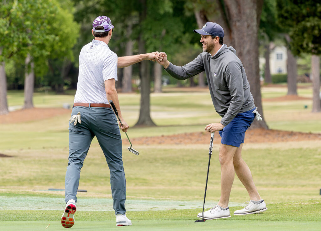 Golfers celebrate with a fist bump on the green during the VersAbility golf tournament, surrounded by lush trees and grass