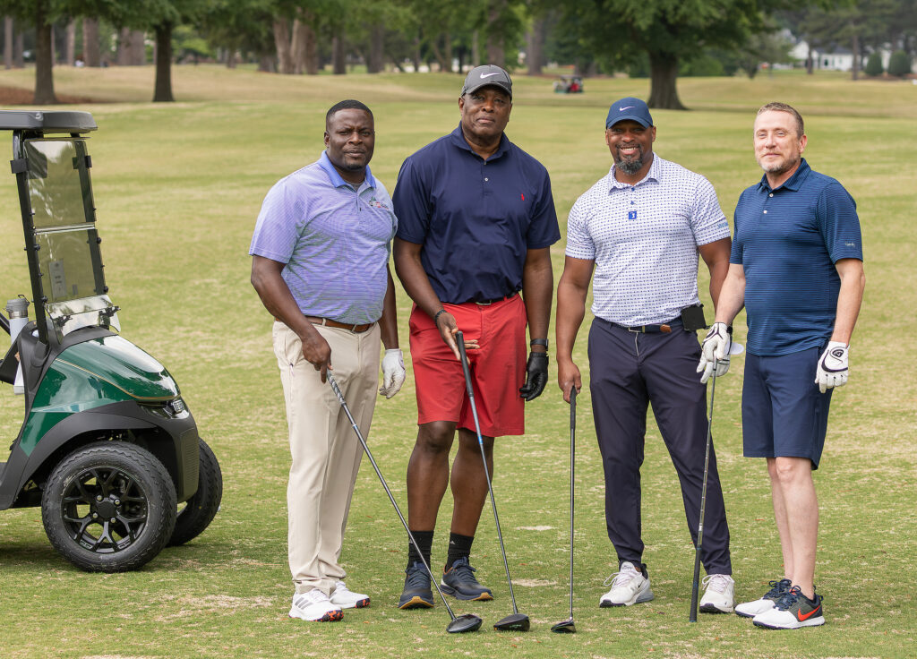 Four men pose with golf clubs beside a cart at the VersAbility golf tournament on a sunny course, ready for a friendly game.
