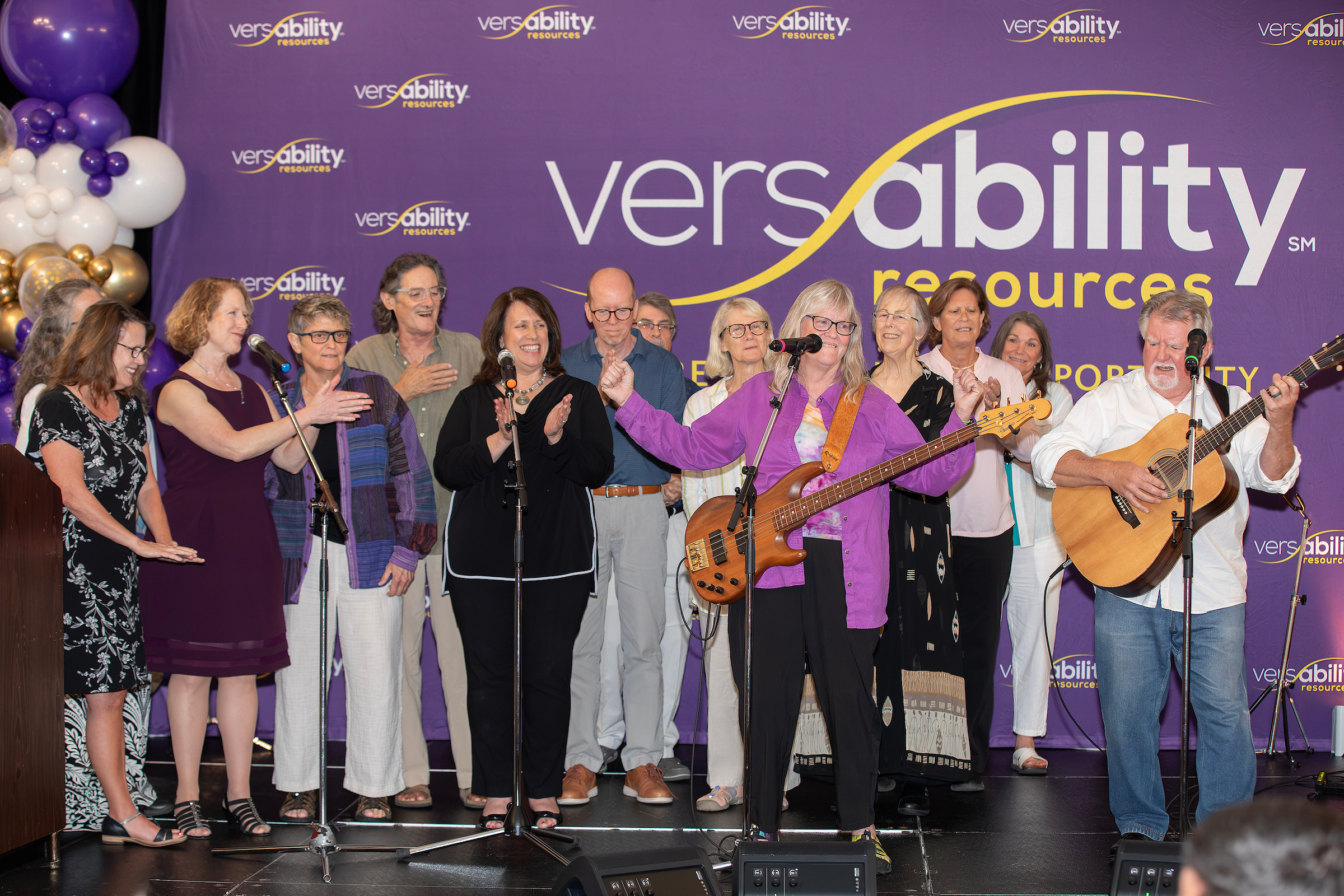 A group of people singing on stage with musical instruments, in front of a purple backdrop featuring the VersAbility Resources logo.