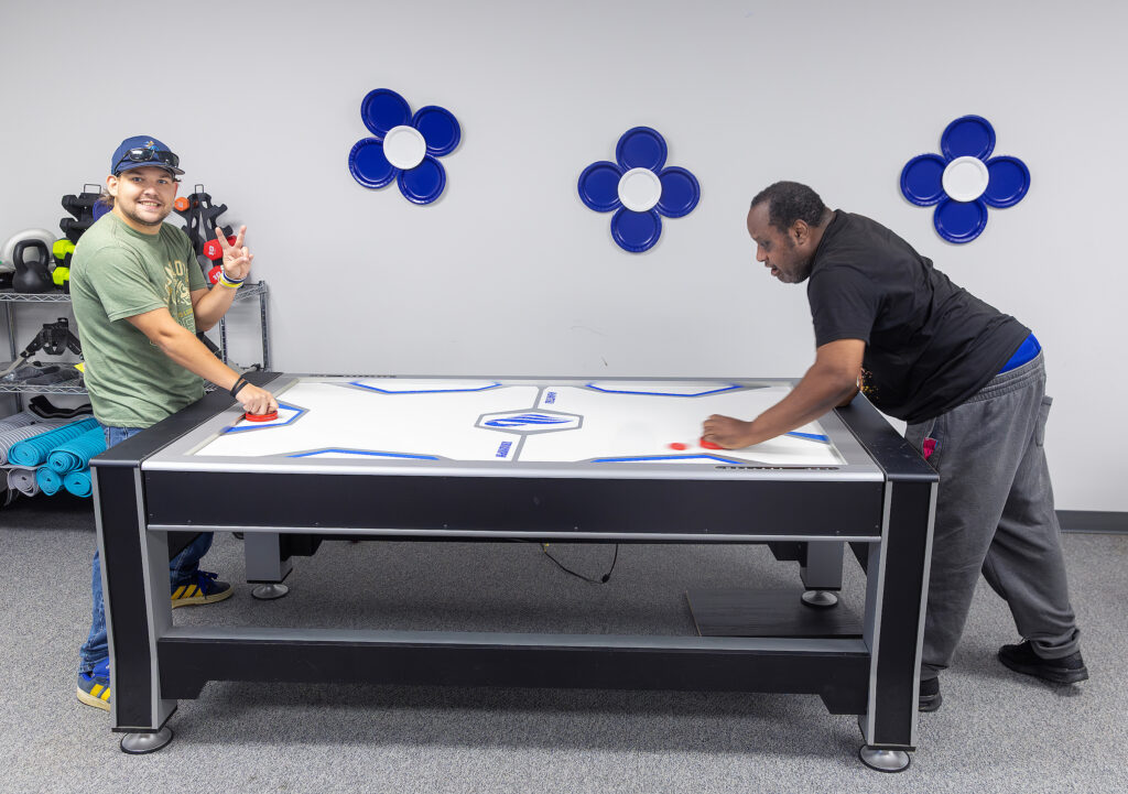 Two men play air hockey in a room with blue wall decor and exercise equipment, enjoying a friendly game.