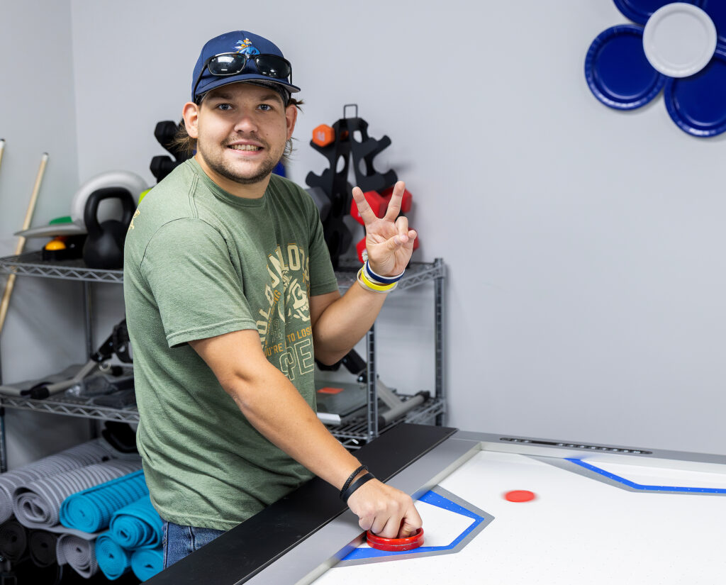 Man in green shirt playing air hockey, smiling, and making a peace sign in a room with shelves and sports equipment.