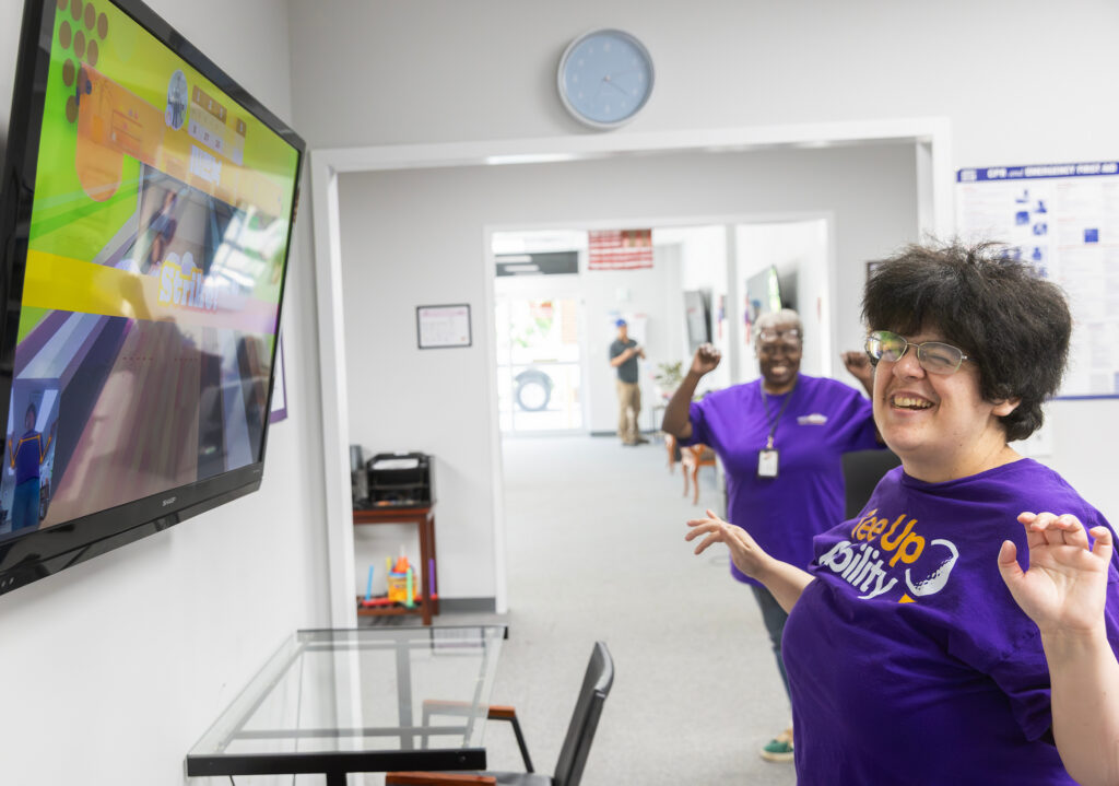 Two people in purple shirts joyfully play a video game in a bright room, celebrating a strike shown on the wall-mounted screen.