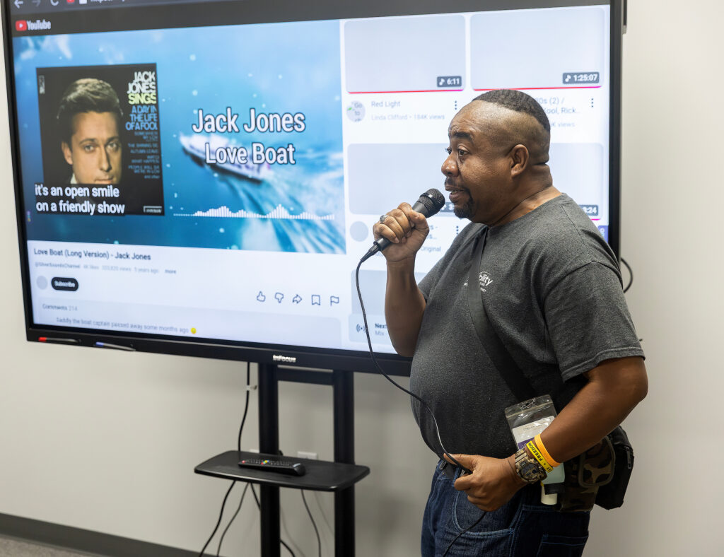 Man sings into a microphone beside a screen displaying "Jack Jones Love Boat" video on YouTube in a casual setting.