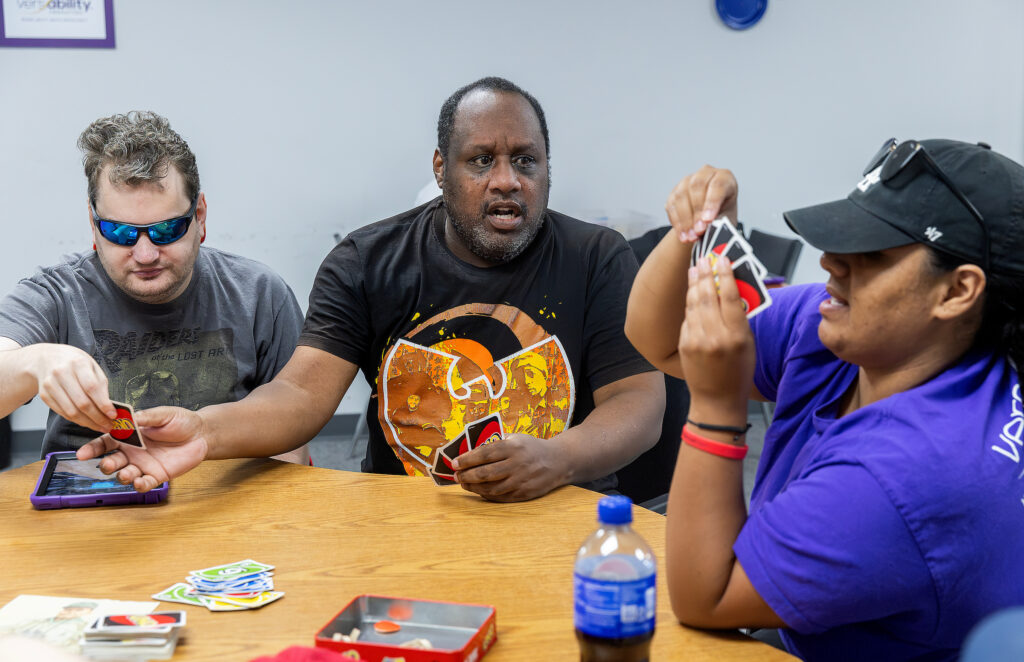 Three people intensely play a card game at a table, with colorful cards and a drink bottle visible.
