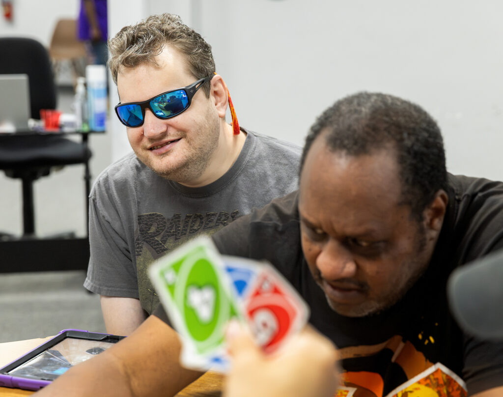 Man in sunglasses plays cards at a table with a tablet. Others in the room engage in activities.