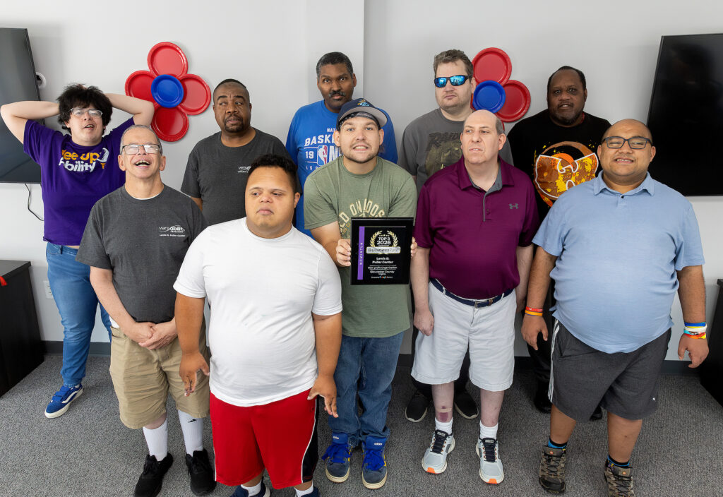 A group of ten people stand together, smiling, with one holding a plaque. Red and blue wall decor is visible in the background.