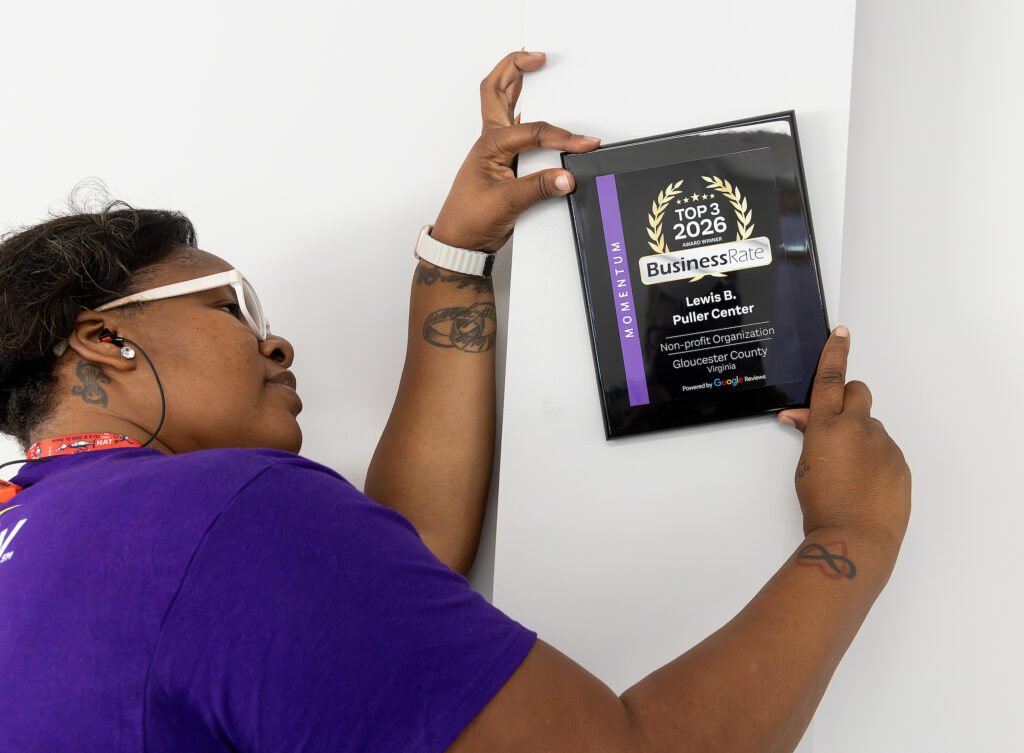 Person in purple shirt hangs a BusinessRate award plaque for Lewis B. Puller Center on a white wall.