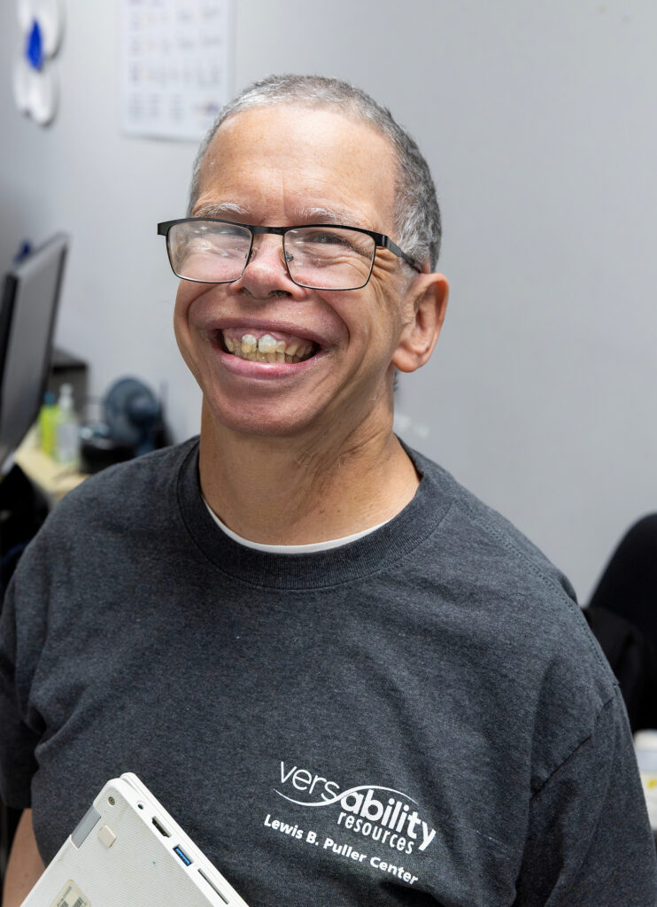 Smiling man in glasses and VersAbility Resources shirt holds a device, standing in an office with computers in the background.