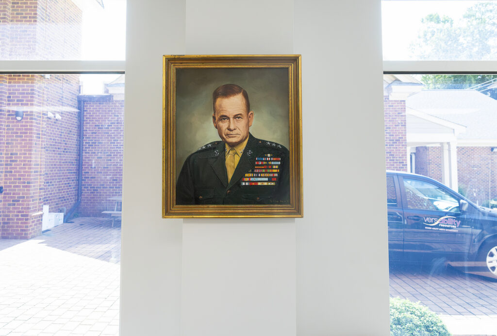 A framed portrait of a military officer with medals hangs on a white wall, with a brick courtyard visible through the window.
