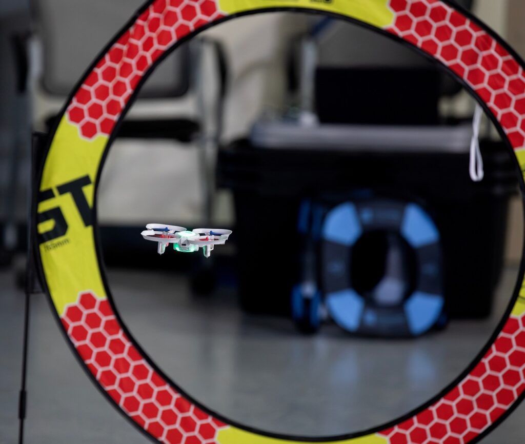 A small drone flies through a circular ring with a yellow and red honeycomb pattern in an indoor setting. There are blurred objects and equipment in the background.