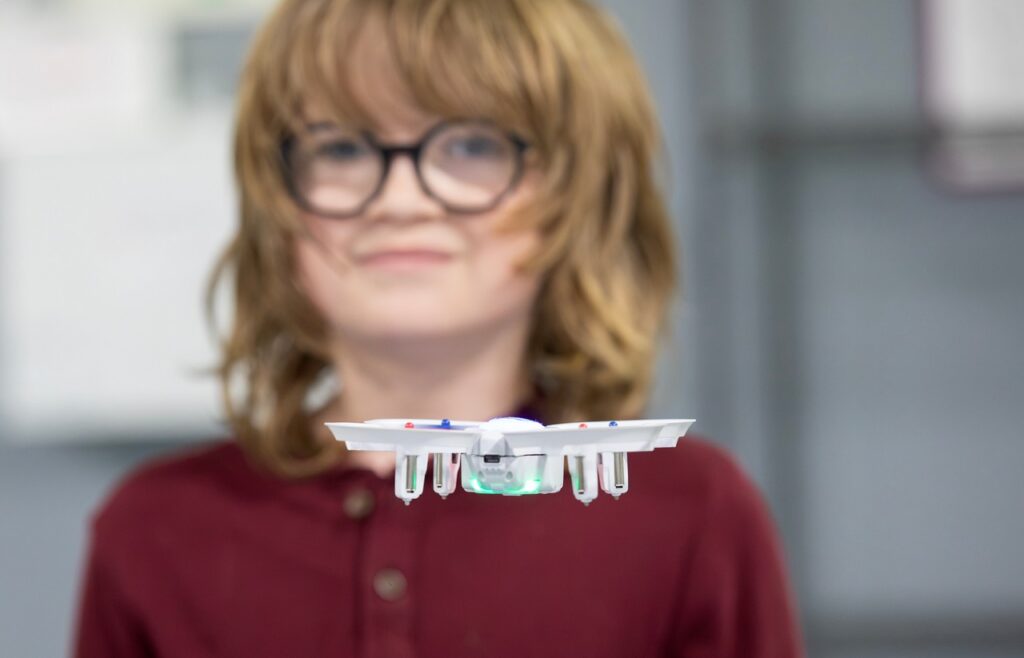 A child with long hair and glasses smiles while watching a small white drone fly in front of them indoors.