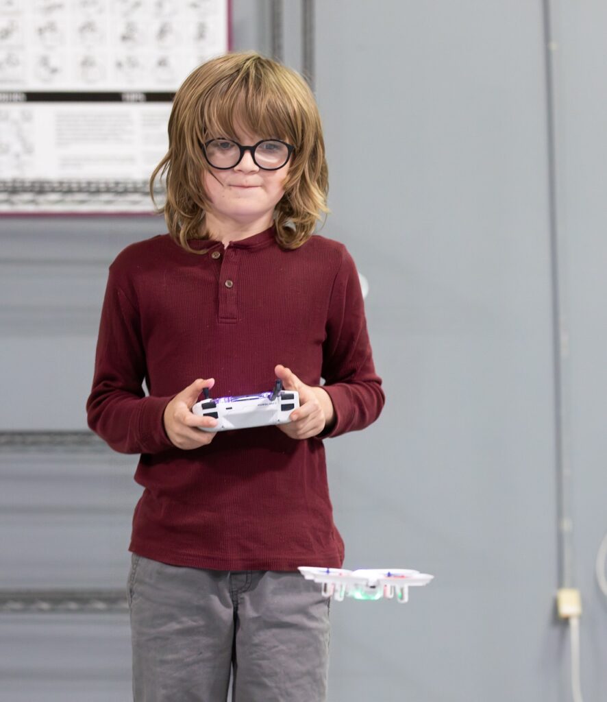 A child wearing glasses and a maroon long-sleeve shirt is flying a small white drone indoors using a remote controller.