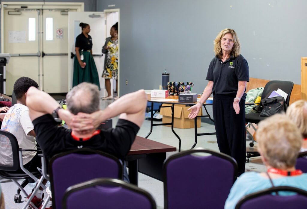 A woman stands and speaks to a seated group in a classroom. Two people talk near the open door in the background. Tables, chairs, and various materials are visible in the room.
