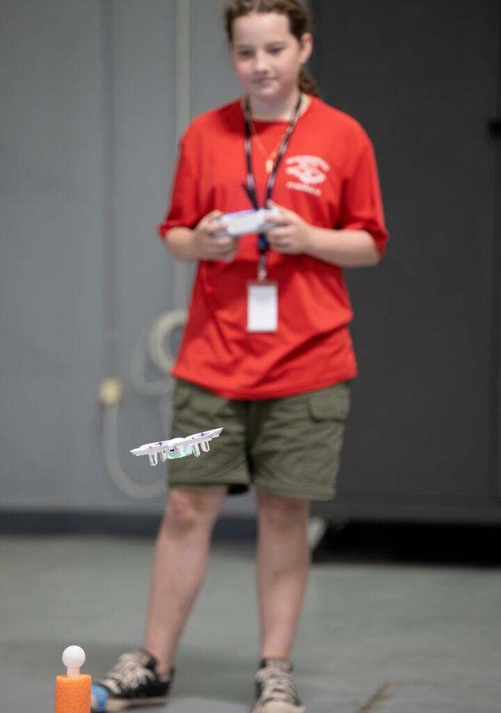 A young person in a red shirt and shorts operates a small drone indoors, focusing as the drone hovers near a foam obstacle with a ball on top.