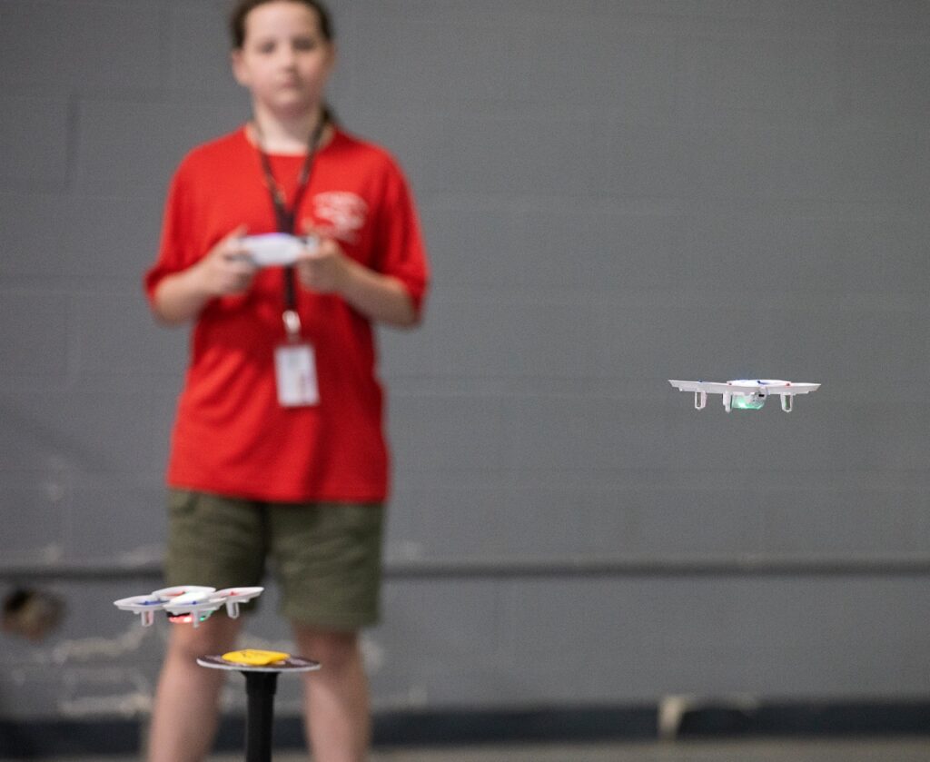 A person in a red shirt and green shorts operates a remote control, flying two small drones indoors. The background is out of focus.