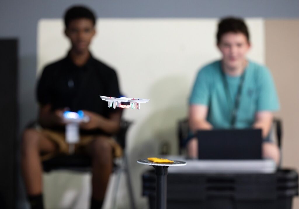Two people sit in the background, one with a laptop, as a small drone hovers in focus between them above a black stand with a yellow object.