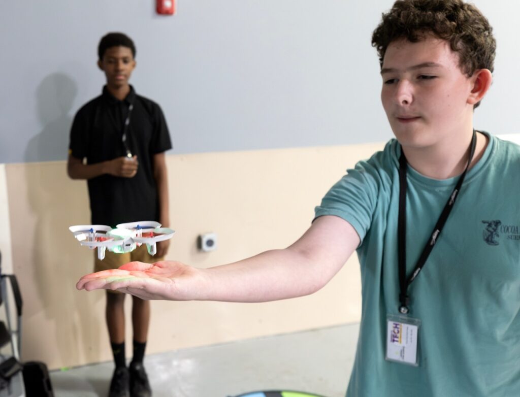 A boy in a teal shirt hovers a small white drone above his hand indoors, while another boy in black stands in the background watching.
