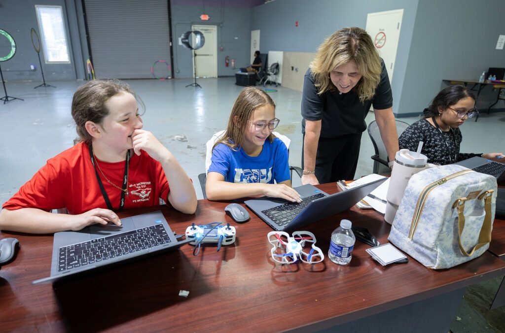 Four people sit at a table with laptops and drones; an adult stands helping three girls who are working and smiling in a tech classroom setting.